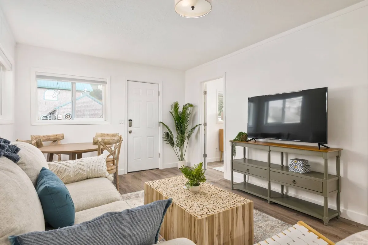 A bright, open-concept living and dining area featuring a comfortable sofa with patterned cushions, a modern coffee table, a compact TV unit, and a dining table with chairs. Lush indoor plants and cheerful decor enhance the welcoming, beachy atmosphere.