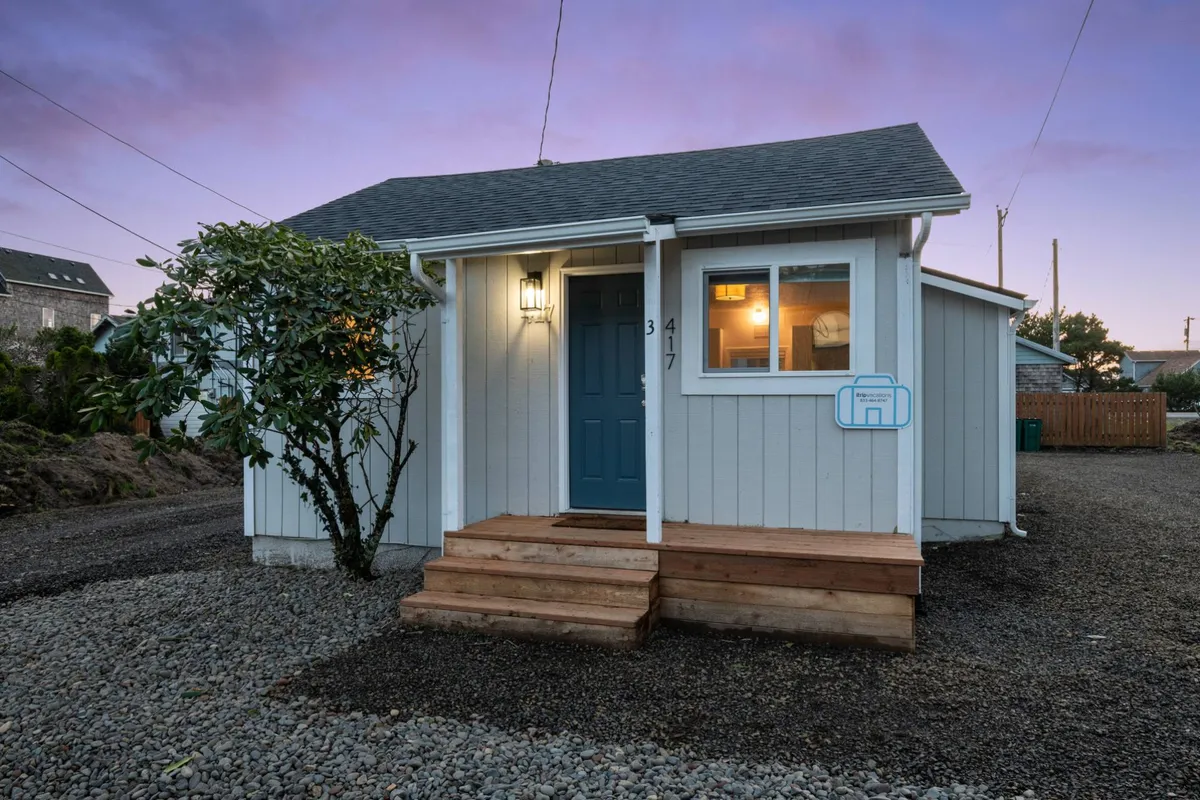 This image showcases the inviting blue front door of a cottage, with wooden steps leading up and lush landscaping surrounding the entrance. The porch area suggests a welcoming space to relax and take in the neighborhood atmosphere.
