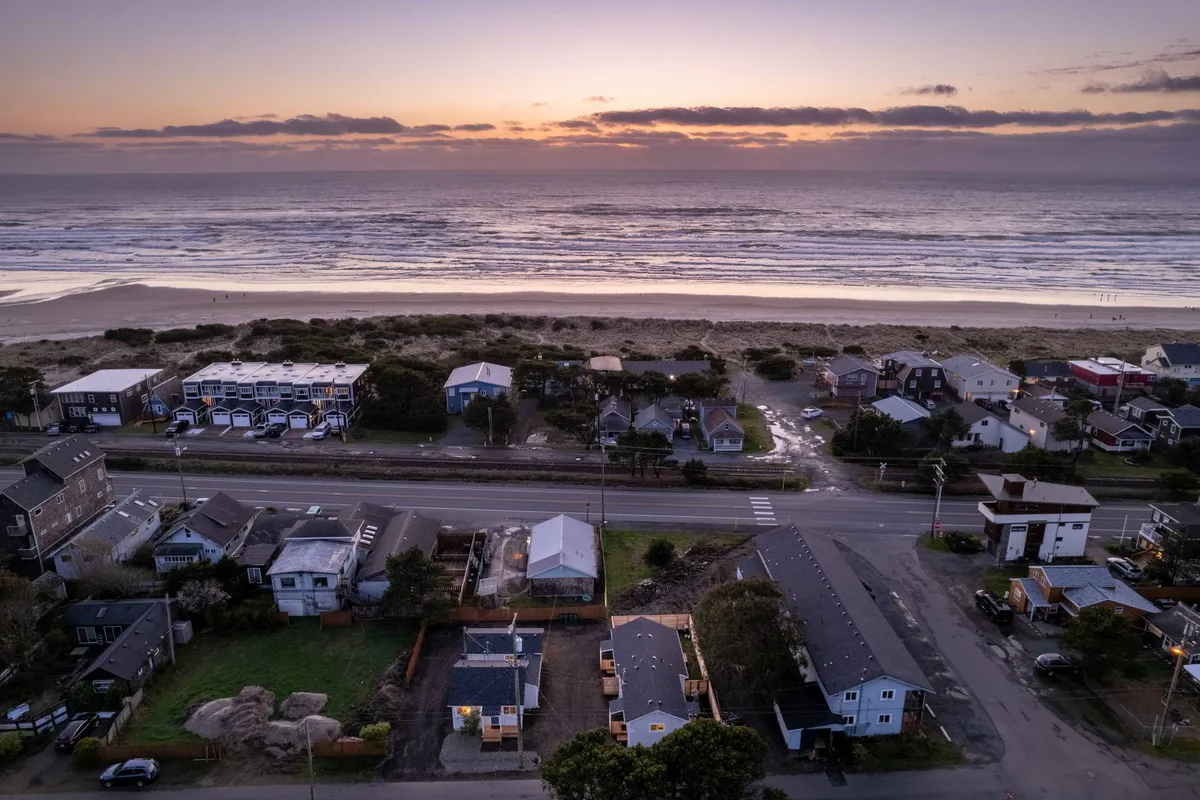 A tranquil scene featuring soft ocean waves, a colorful sunset sky, beachfront houses, and dune grass along the shore. The view suggests a peaceful, welcoming atmosphere, perfect for romantic sunset walks or connecting with neighbors over beach activities.
