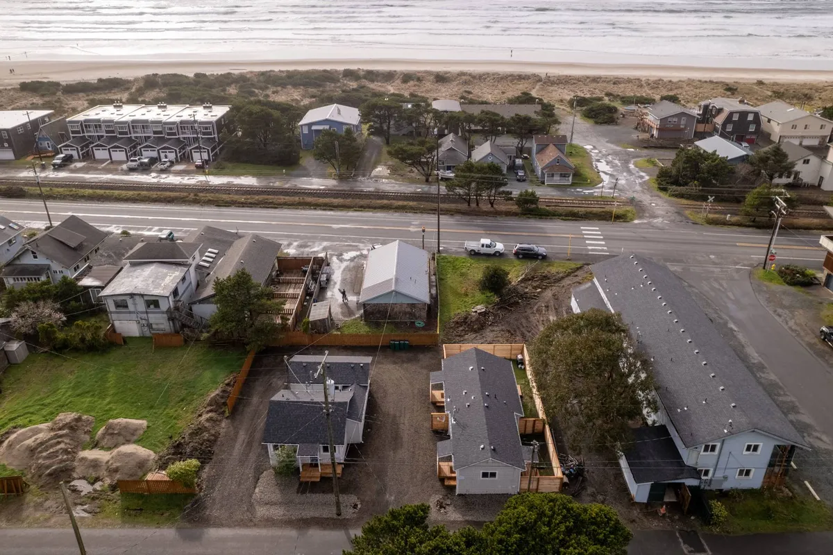 A scenic view of beachfront properties lining the road that leads directly to the sandy expanse of Rockaway Beach. Gentle ocean waves roll in under a pastel sky, emphasizing easy access to the shoreline and the relaxed coastal lifestyle. The image evokes a sense of proximity between cozy accommodations and the inviting beach just steps away.