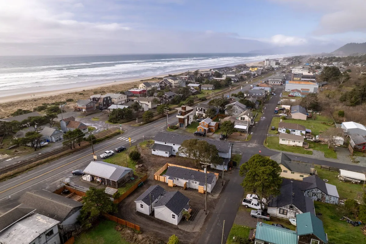 A broad view of the coastline with the ocean stretching beside the sandy beach, houses nearby, and a road lined with trees. The scene captures a variety of beach activities, from walking and biking to picnicking and sandcastle building, conveying the diversity of adventures available just outside the cottage.