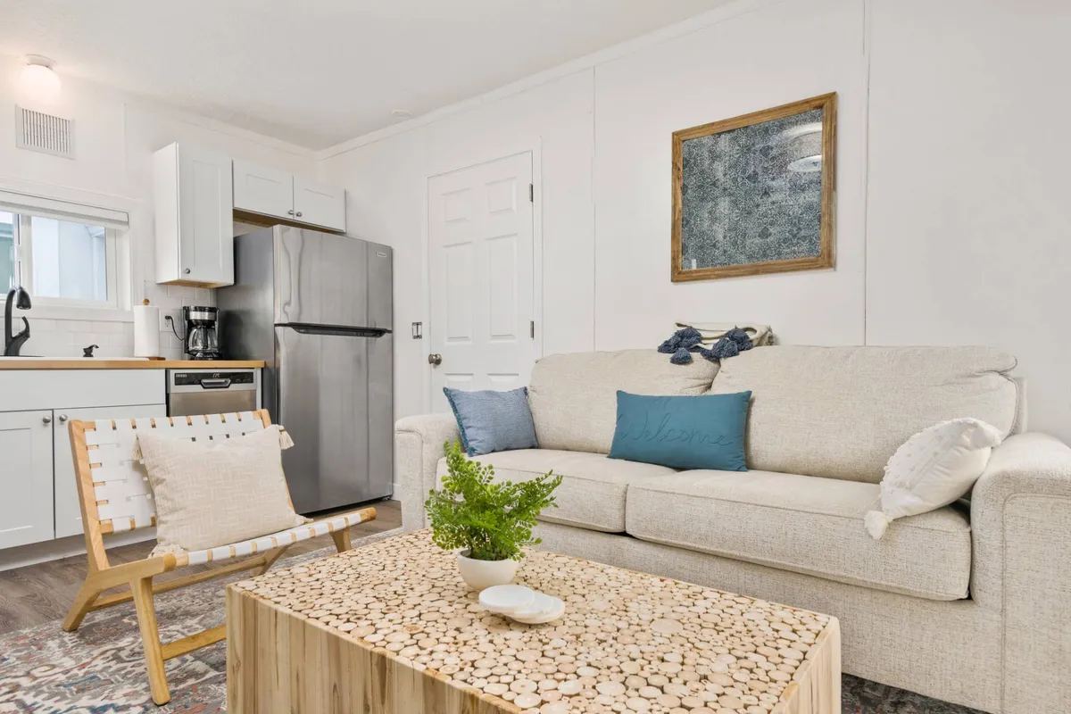 A cozy interior scene centers on a comfortable sofa and coffee table with the kitchen visible in the background. The space feels welcoming and uncluttered, ideal for relaxing with a pet after a day of adventures.