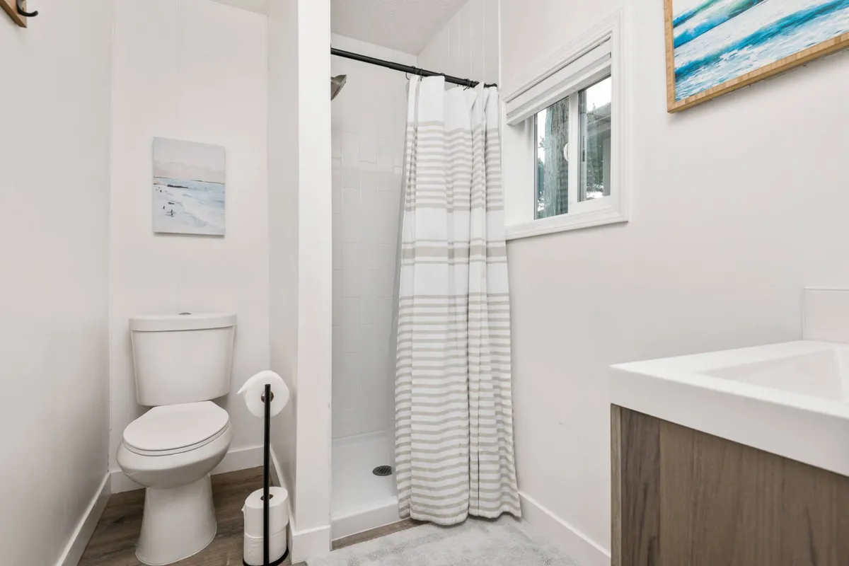 A calm and modern ensuite bathroom with a toilet, walk-in shower, sink, and tasteful artwork. The design is clean and updated, creating an inviting space for personal routines and refreshment after a day at the coast.